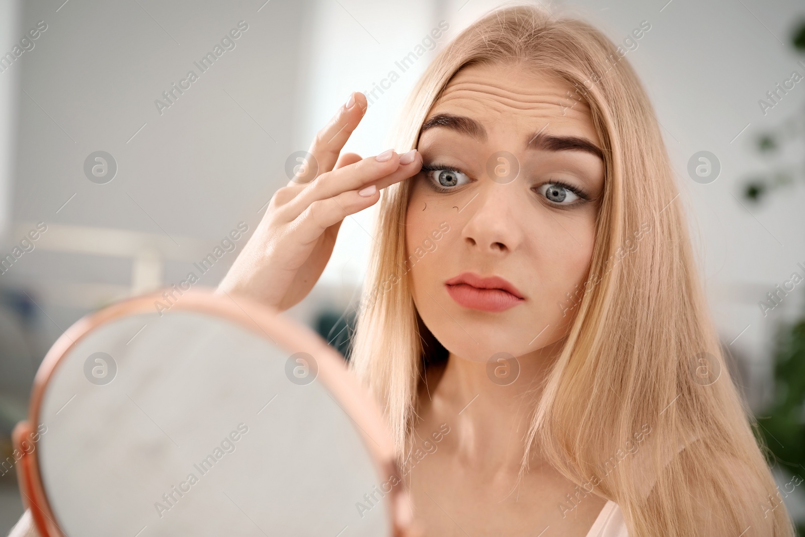 Photo of Young woman with eyelash loss problem looking in mirror indoors