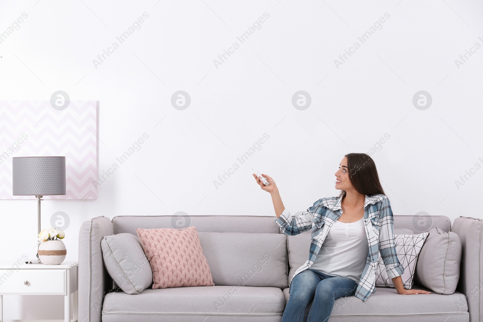 Photo of Young woman switching on air conditioner while sitting on sofa near white wall