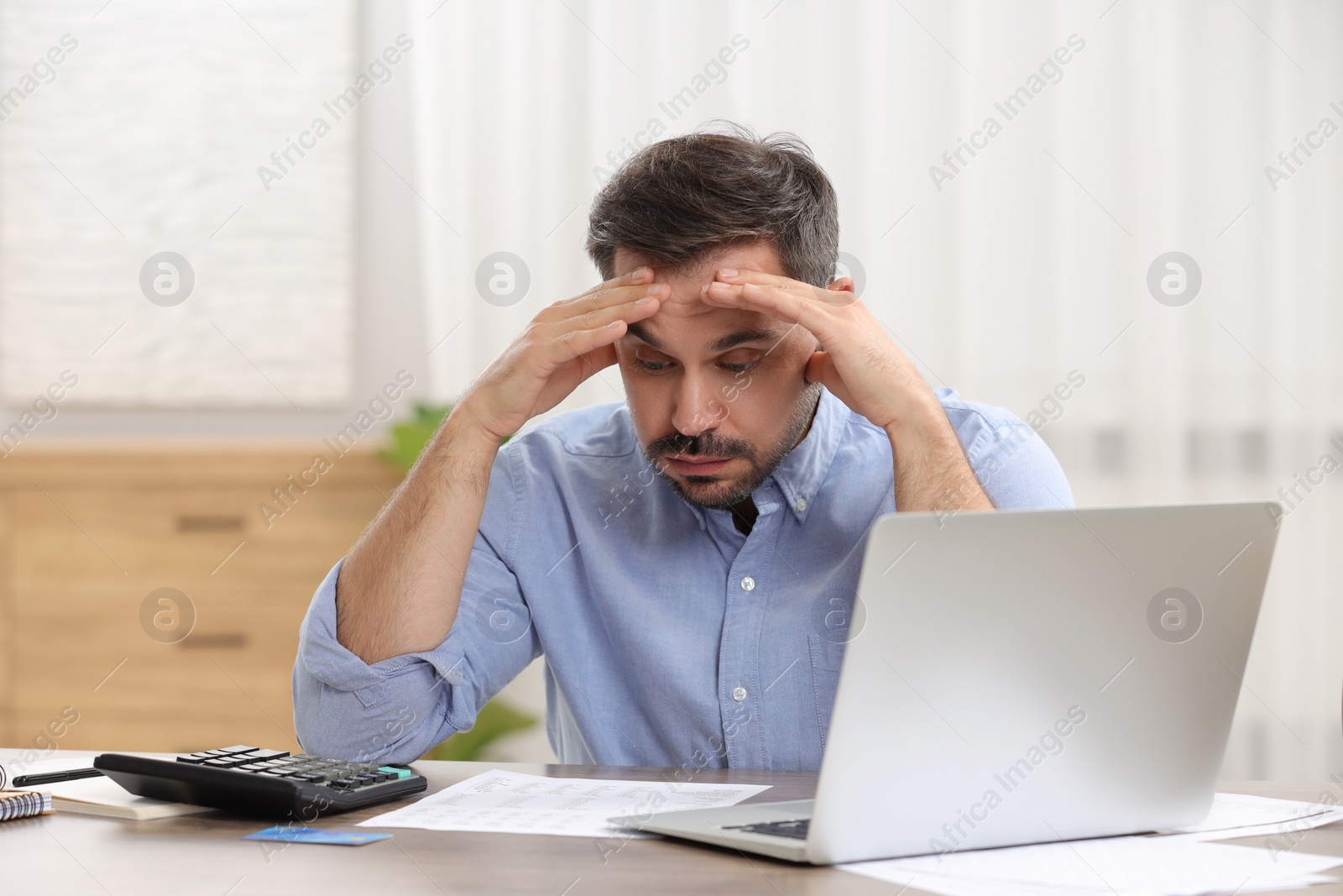 Photo of Confused man with debt notification, laptop and credit card planning budget at table in room. Financial problem