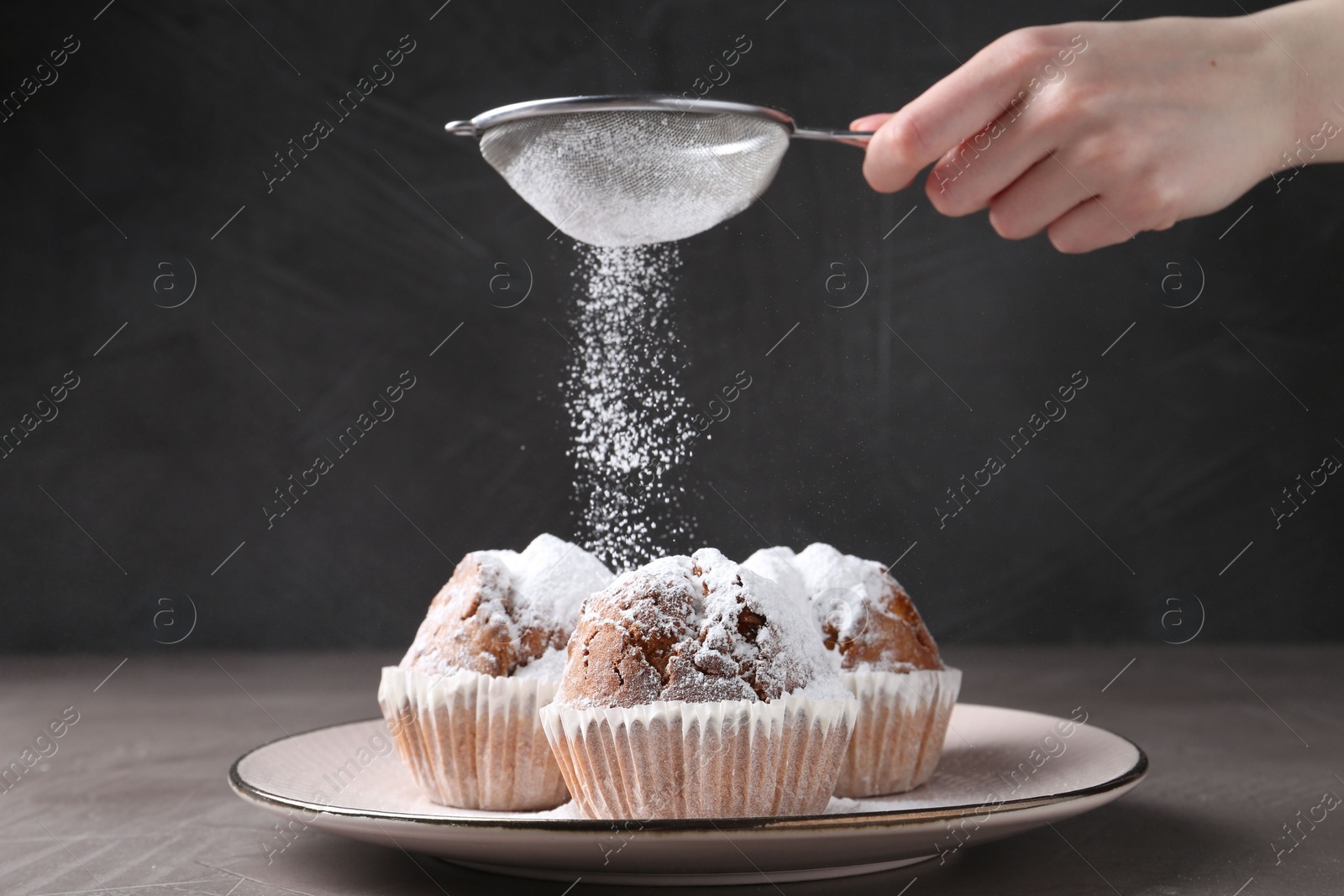 Photo of Woman with sieve sprinkling powdered sugar onto muffins at grey textured table, closeup