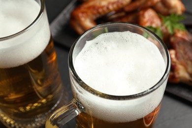 Photo of Mugs with delicious beer and baked chicken wings on table, closeup