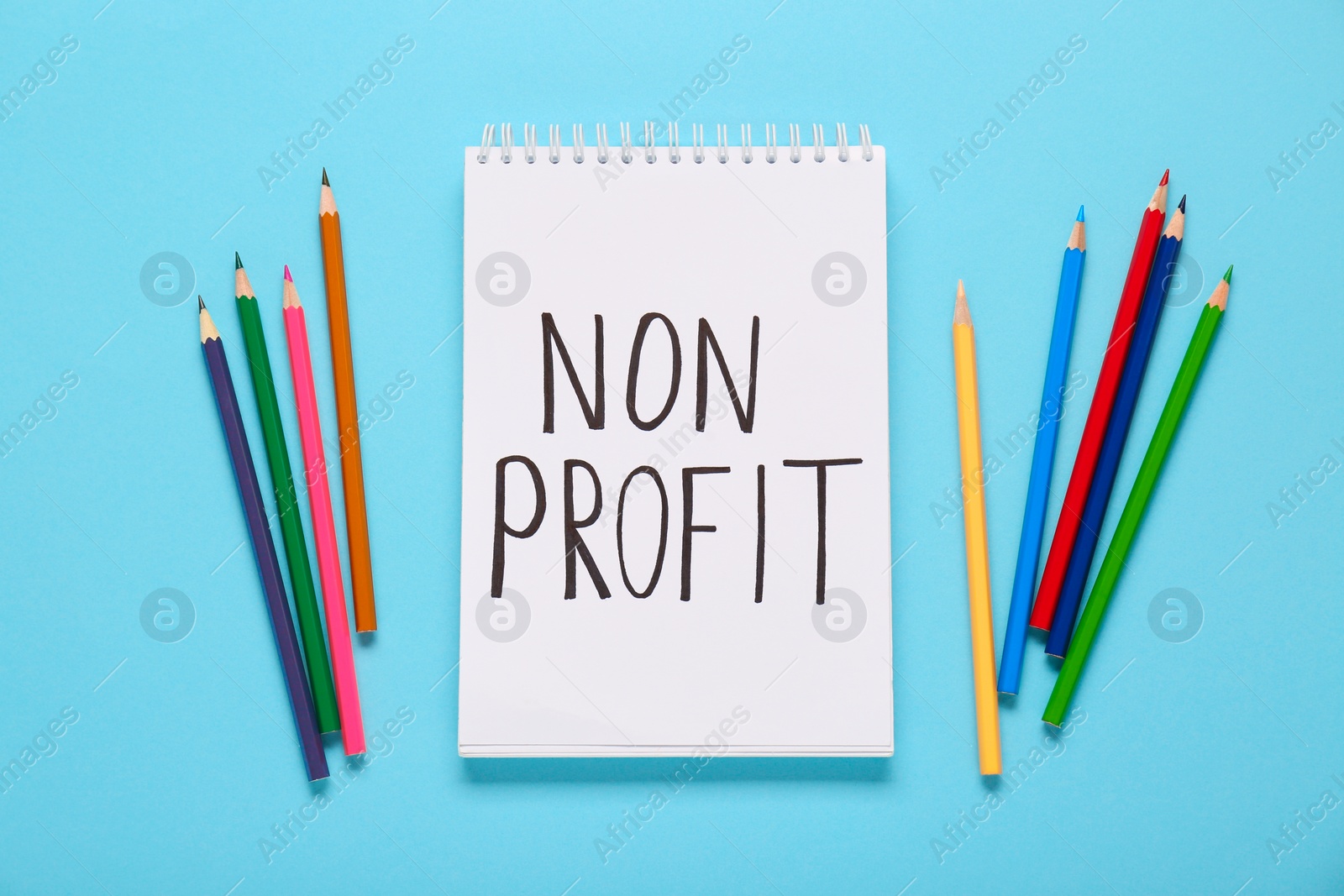 Photo of Notebook with phrase Non Profit and colorful pencils on light blue background, flat lay