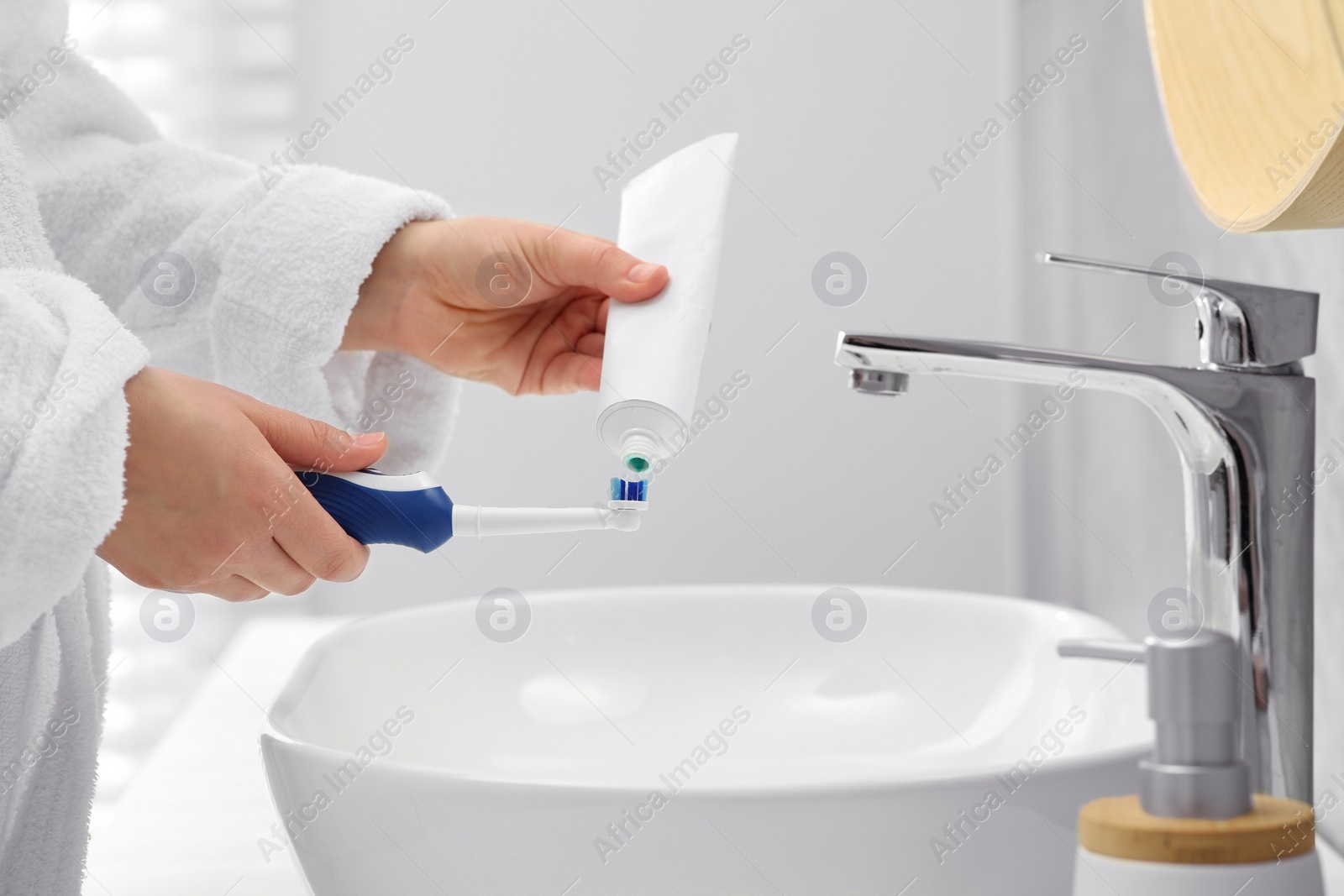 Photo of Woman squeezing toothpaste from tube onto electric toothbrush above sink in bathroom, closeup