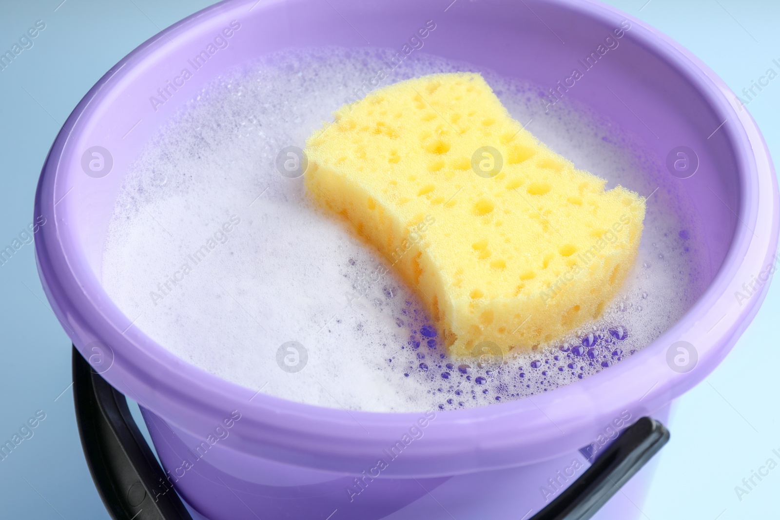 Photo of Bucket with foam and sponge on light blue background, closeup