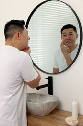 Photo of Handsome man applying cream onto his face near mirror in bathroom