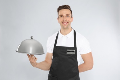 Handsome waiter holding metal tray with lid on light background