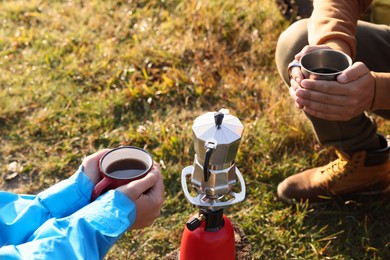 Photo of Tourists with mugs of coffee sitting around camping burner outdoors, closeup