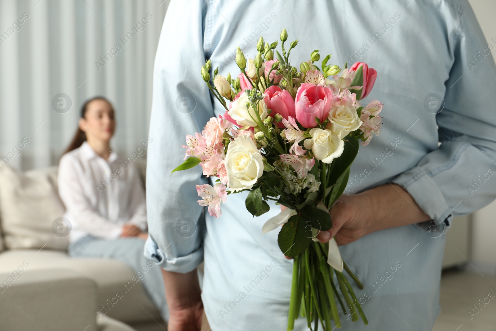 Photo of Man hiding bouquet of flowers for his beloved woman indoors, closeup