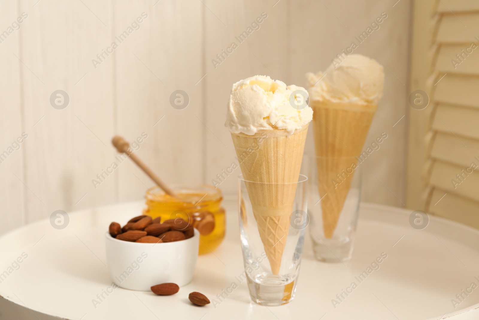 Photo of Delicious vanilla ice cream in wafer cones with honey and almonds on white table indoors