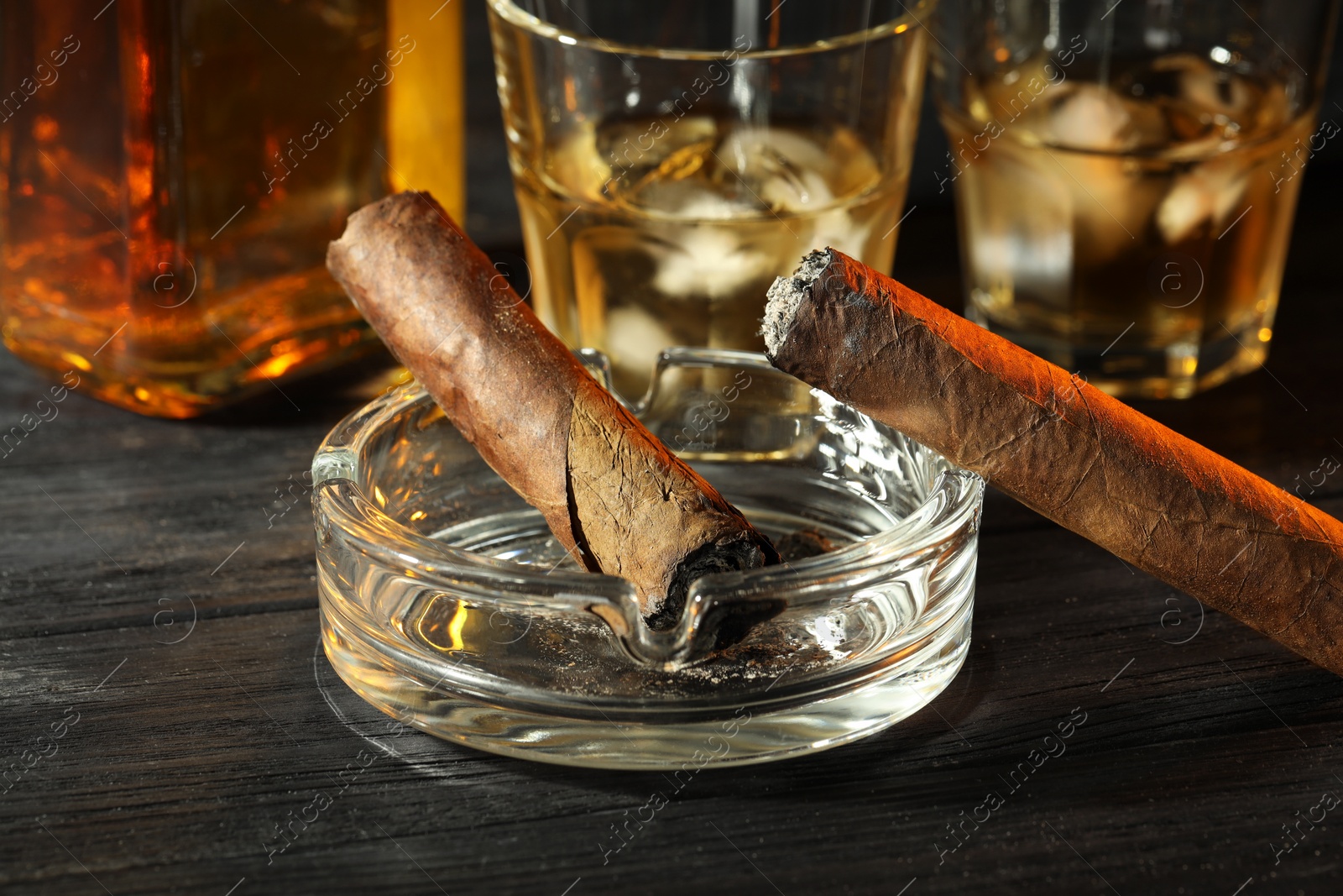 Photo of Cigars, ashtray and whiskey with ice cubes on black wooden table, closeup