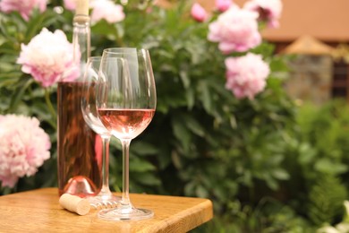 Photo of Glasses and bottle with rose wine on wooden table near beautiful peonies, space for text