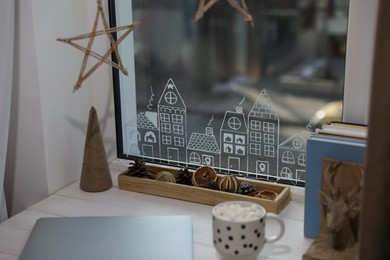 Laptop and hot drink on white wooden table near window with beautiful drawing at home. Christmas decor