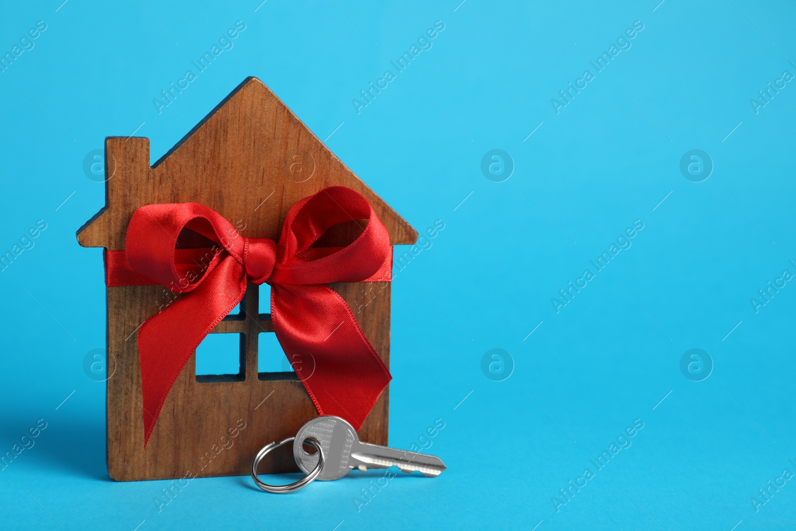 Photo of Wooden house model with red bow and key on light blue background, space for text. Housewarming party