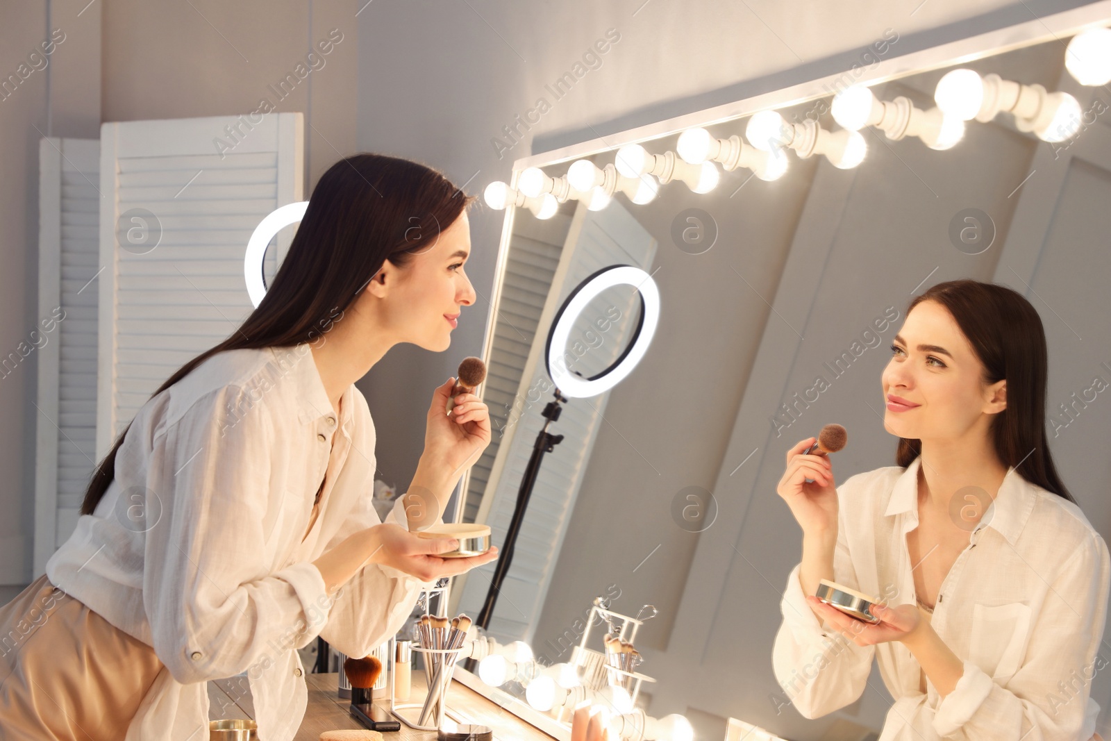 Photo of Young woman applying make up near illuminated mirror indoors