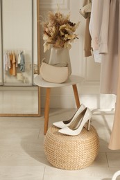 Elegant women's shoes on wicker pouf near stylish clothes in dressing room. Interior design