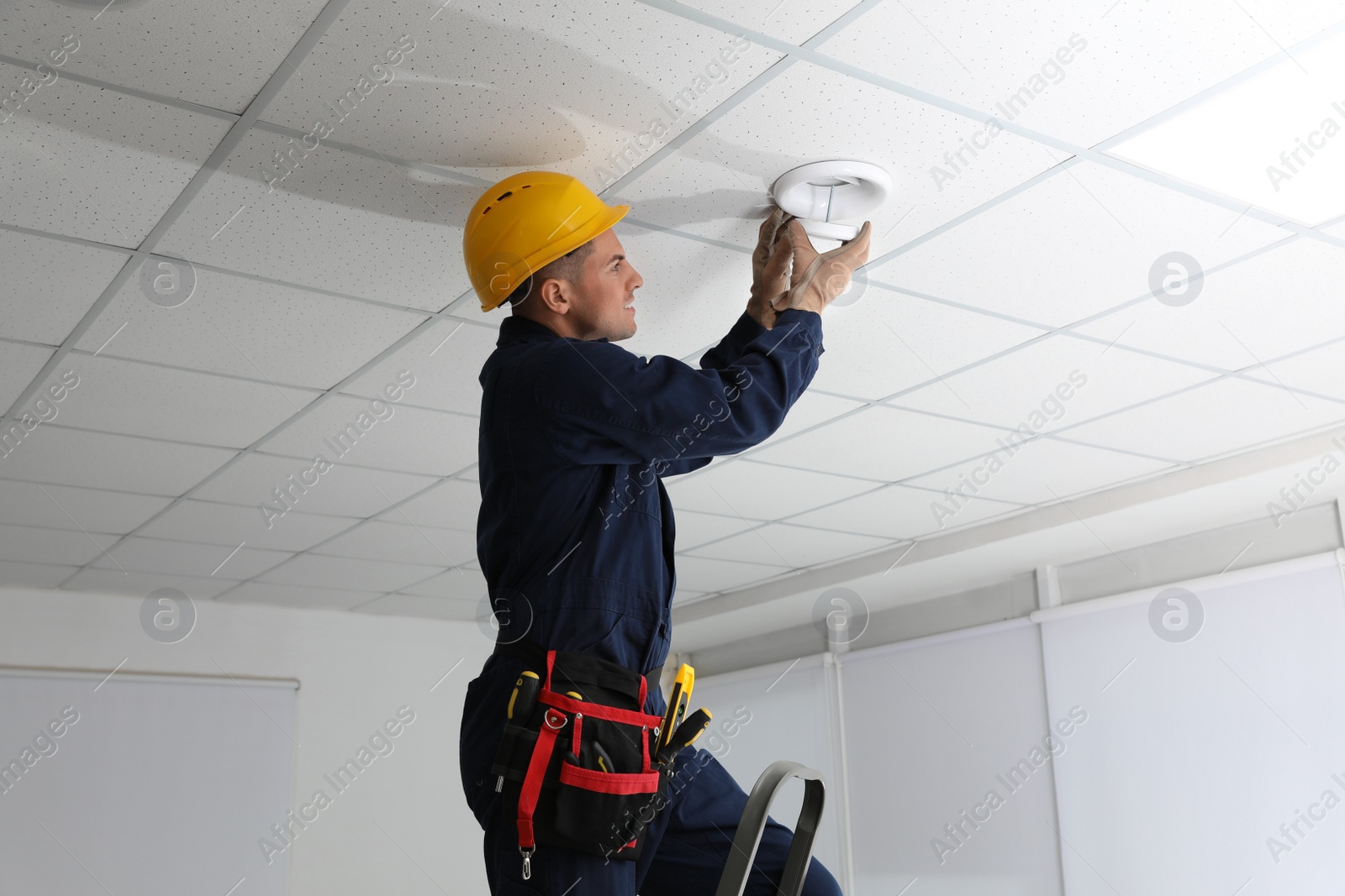 Photo of Electrician in uniform repairing ceiling lamp indoors