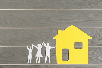 Photo of Paper figures of happy family holding hands and yellow house on grey wooden background, flat lay