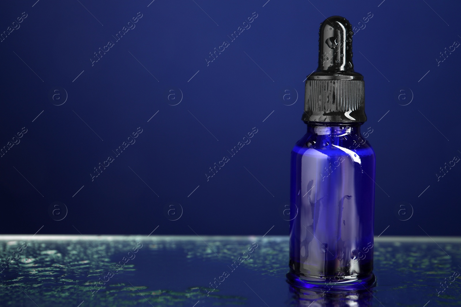 Photo of Bottle of face serum on wet surface against blue background, closeup. Space for text