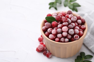 Photo of Frozen red cranberries in bowl and green leaves on white table, closeup. Space for text