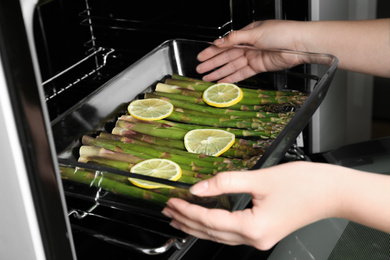 Photo of Woman putting glass baking dish with raw asparagus and lemon slices in oven, closeup