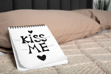 Photo of Notebook with phrase Kiss Me and hearts on bed in room, closeup. Space for text