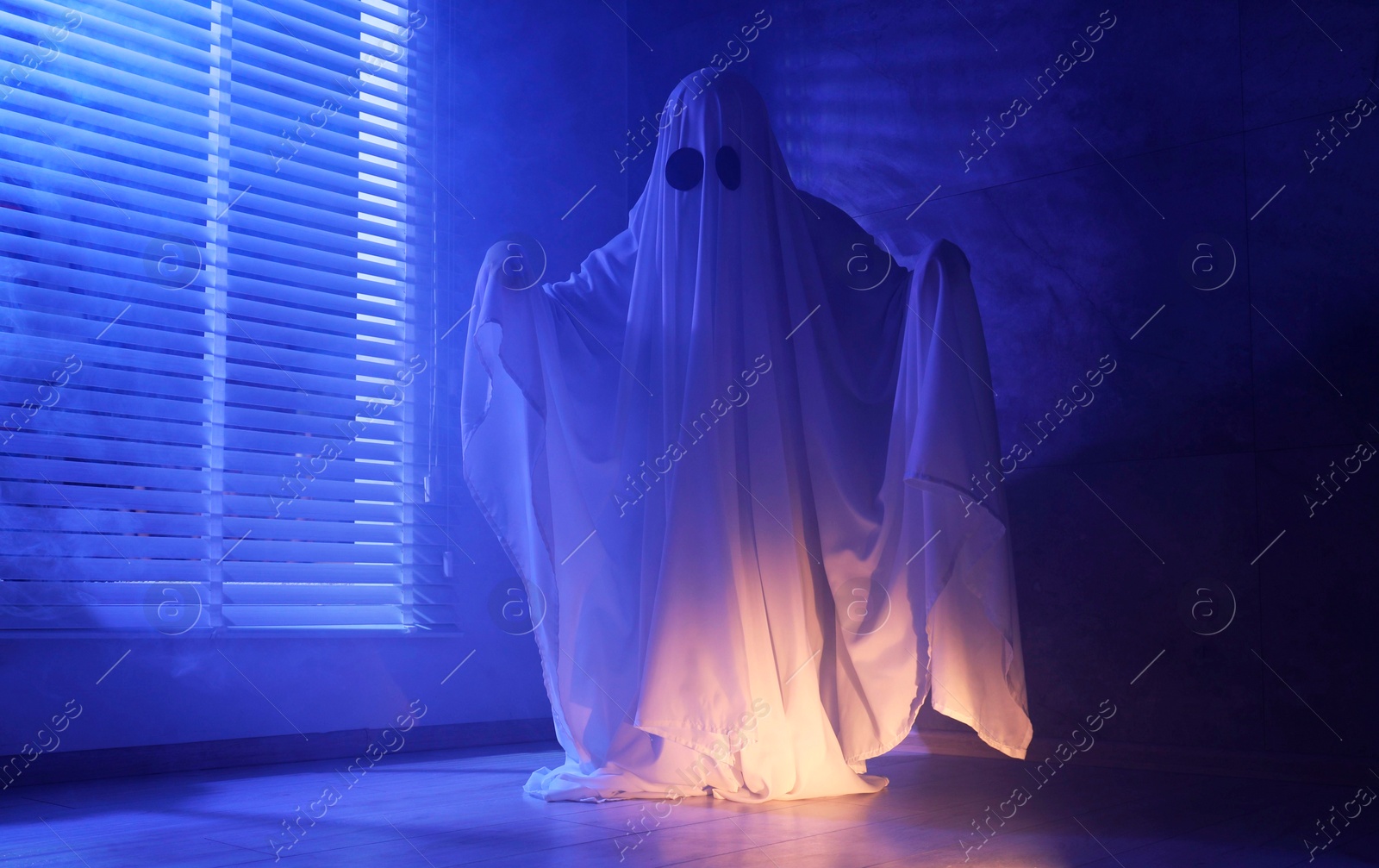 Photo of Creepy ghost. Woman covered with sheet near window in blue light