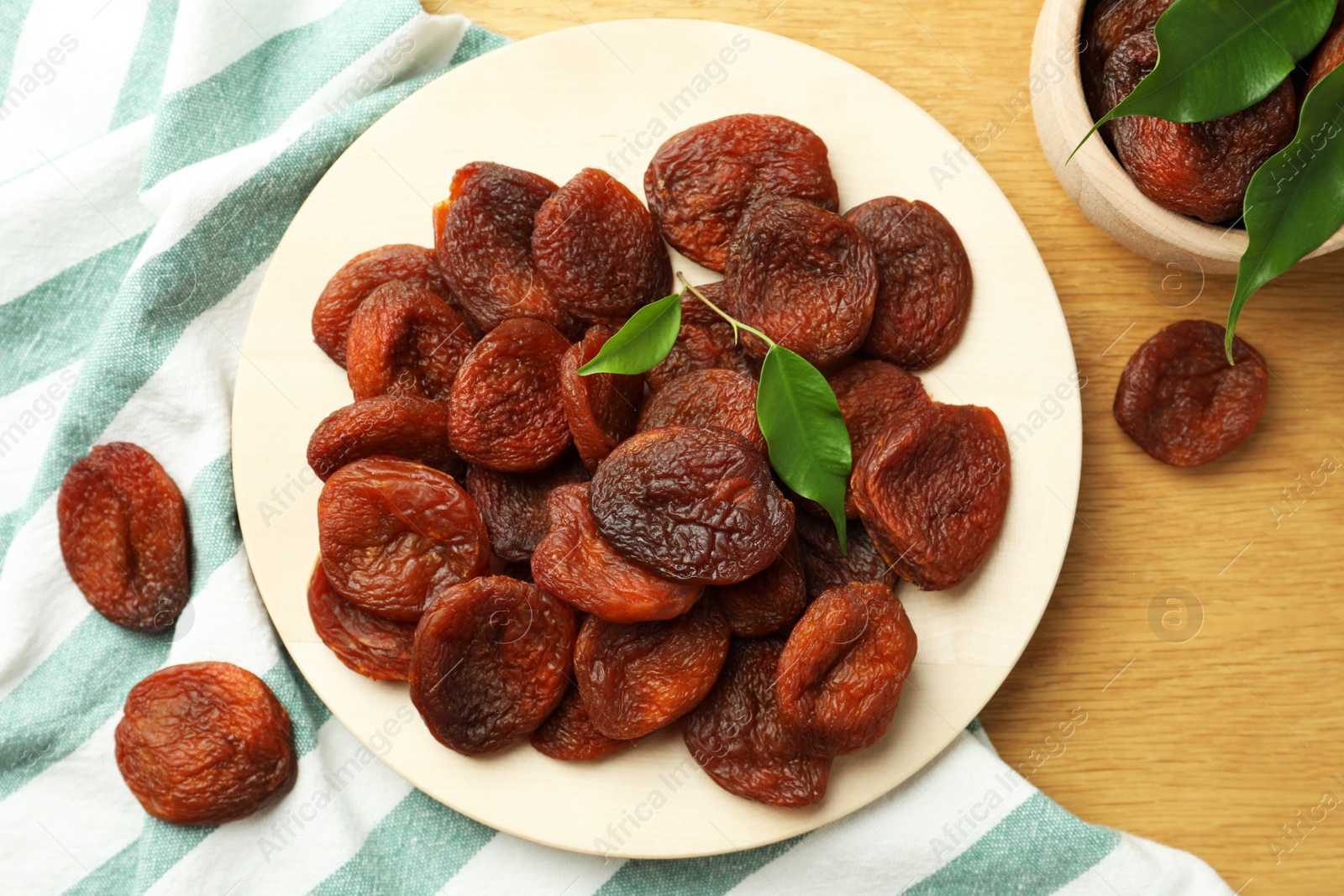 Photo of Tasty apricots and green leaves with plate on wooden table, flat lay. Dried fruits