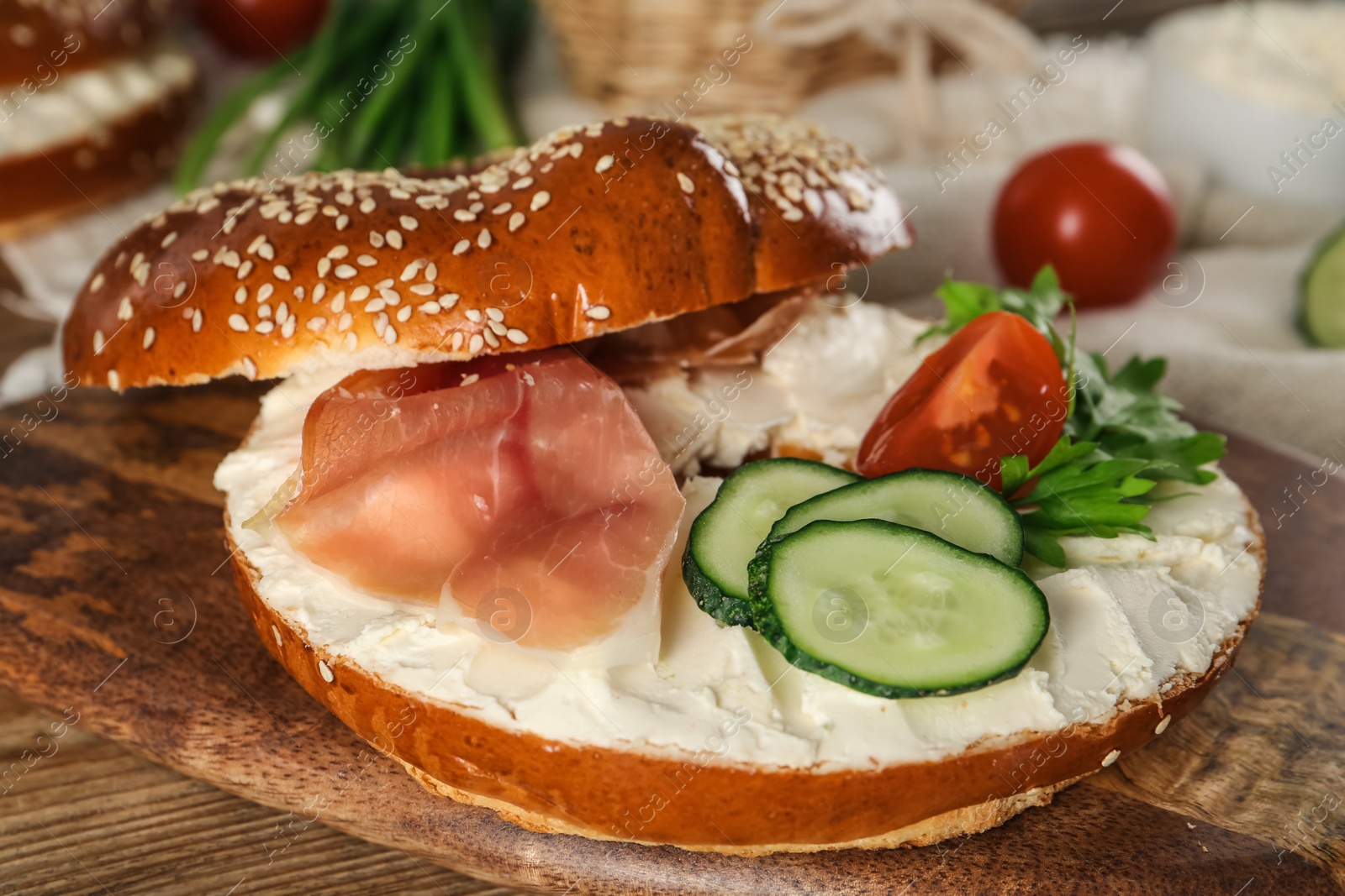 Photo of Delicious bagel with cream cheese, jamon, cucumber, tomato and parsley on wooden board, closeup