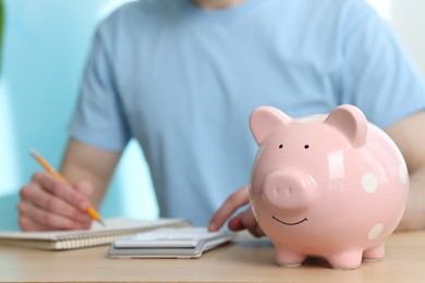 Financial savings. Man writing down notes at wooden table, focus on piggy bank