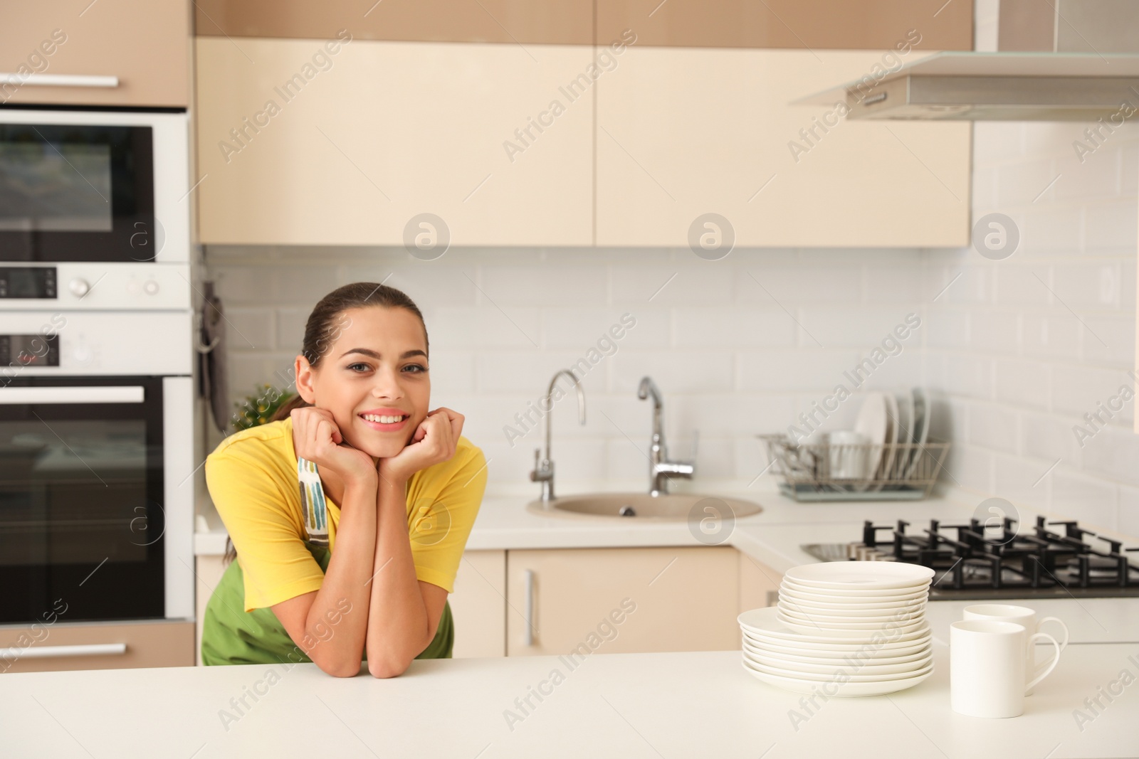 Photo of Beautiful young woman with clean dishes and cups at table in kitchen