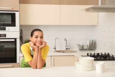 Photo of Beautiful young woman with clean dishes and cups at table in kitchen