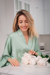 Photo of Pretty young woman in beautiful silk robe with flowers at kitchen table