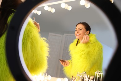 Photo of Young woman applying make up near illuminated mirror indoors, view through ring lamp