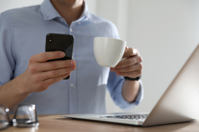 Man with cup of coffee and smartphone at table indoors, closeup