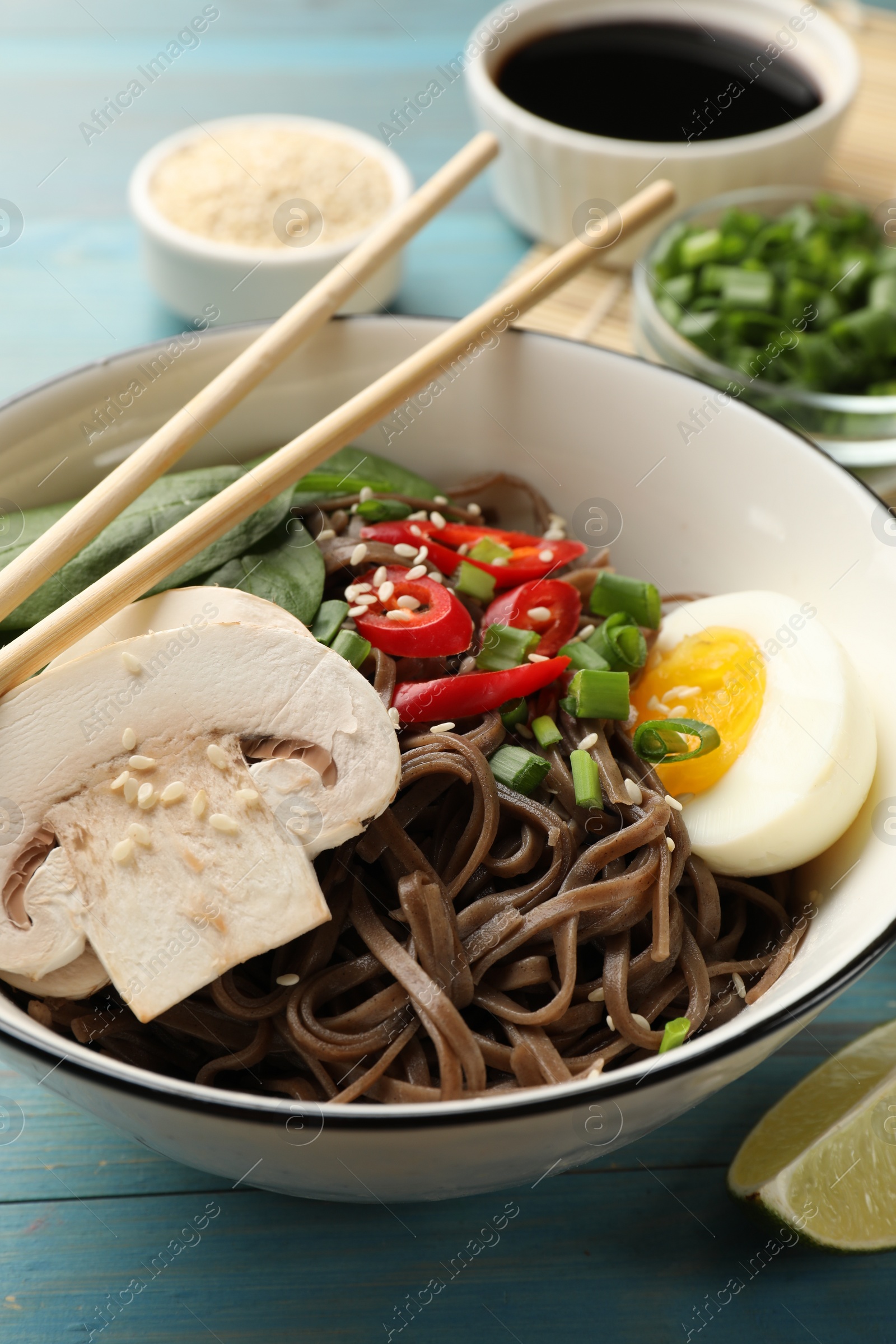 Photo of Tasty buckwheat noodles (soba) with chili pepper, egg, mushrooms and chopsticks on light blue wooden table, closeup