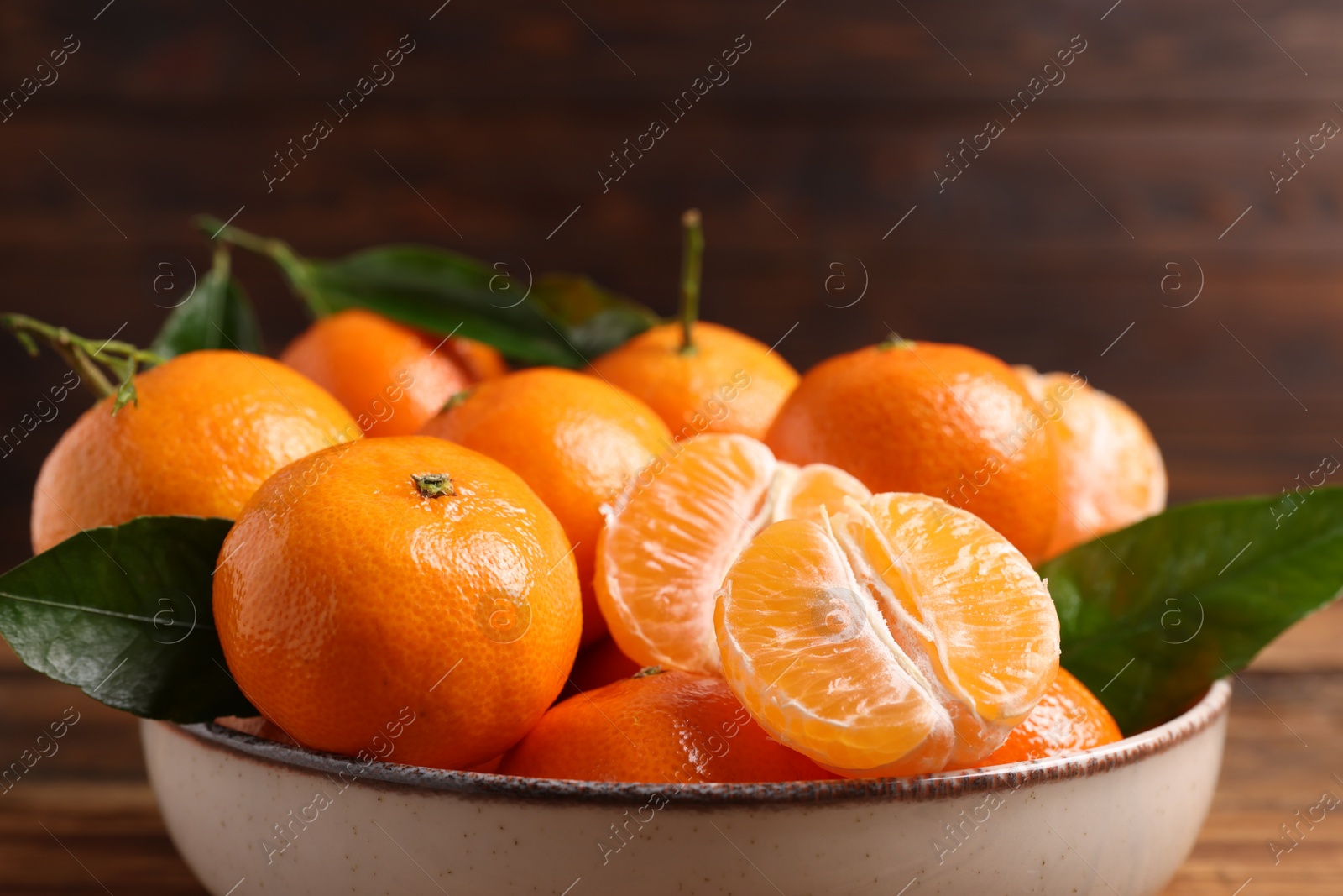 Photo of Fresh tangerines with green leaves in bowl on wooden table, closeup