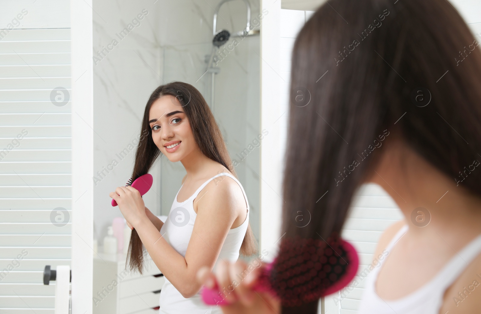 Photo of Beautiful young woman with hair brush looking into mirror in bathroom