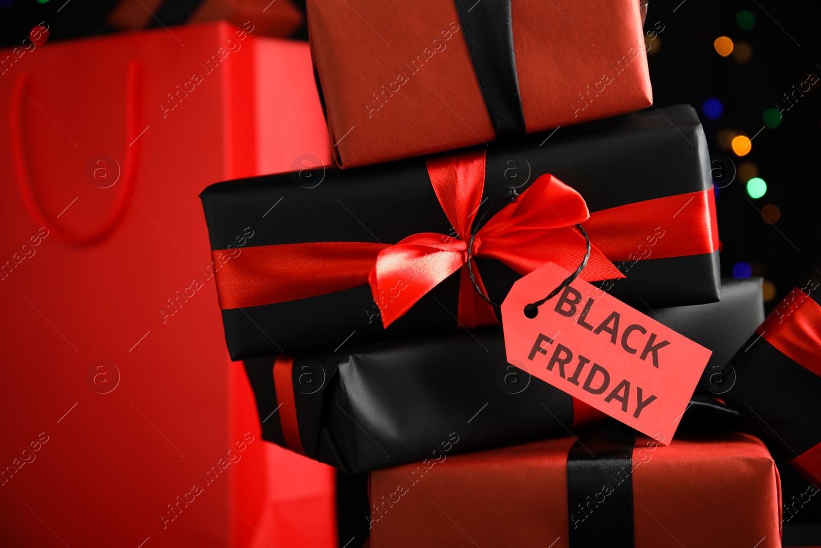Photo of Gift boxes with Black Friday tag and shopping bag against blurred lights, closeup