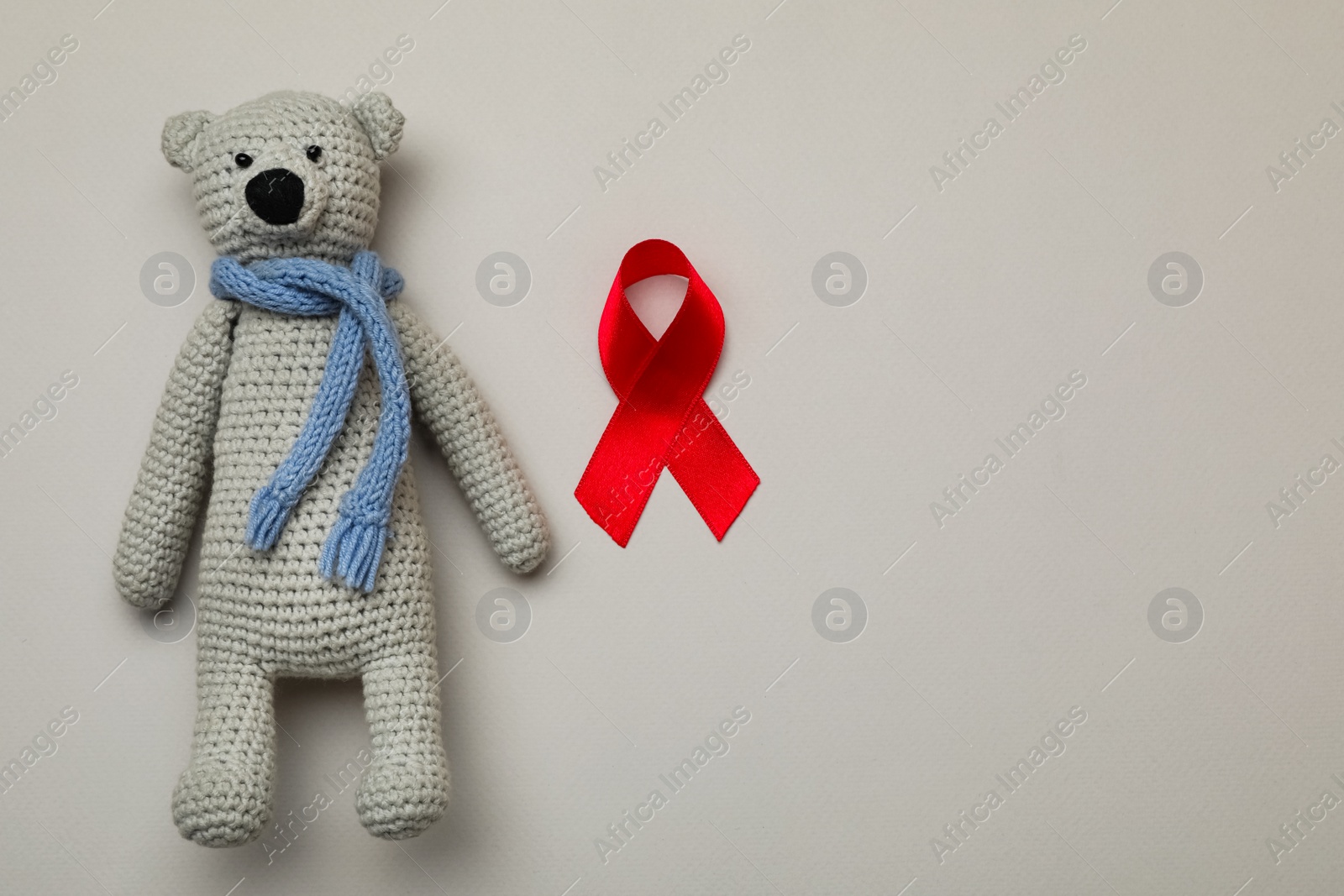 Photo of Cute knitted toy bear and red ribbon on beige background, flat lay with space for text. AIDS disease awareness