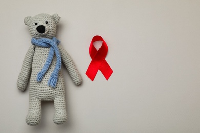 Photo of Cute knitted toy bear and red ribbon on beige background, flat lay with space for text. AIDS disease awareness