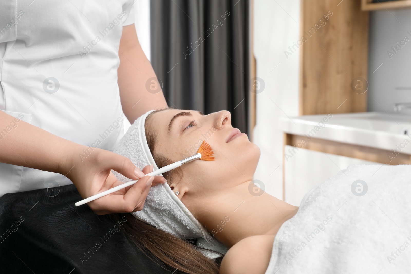 Photo of Cosmetologist with brush applying cosmetic product to client`s face in clinic, closeup