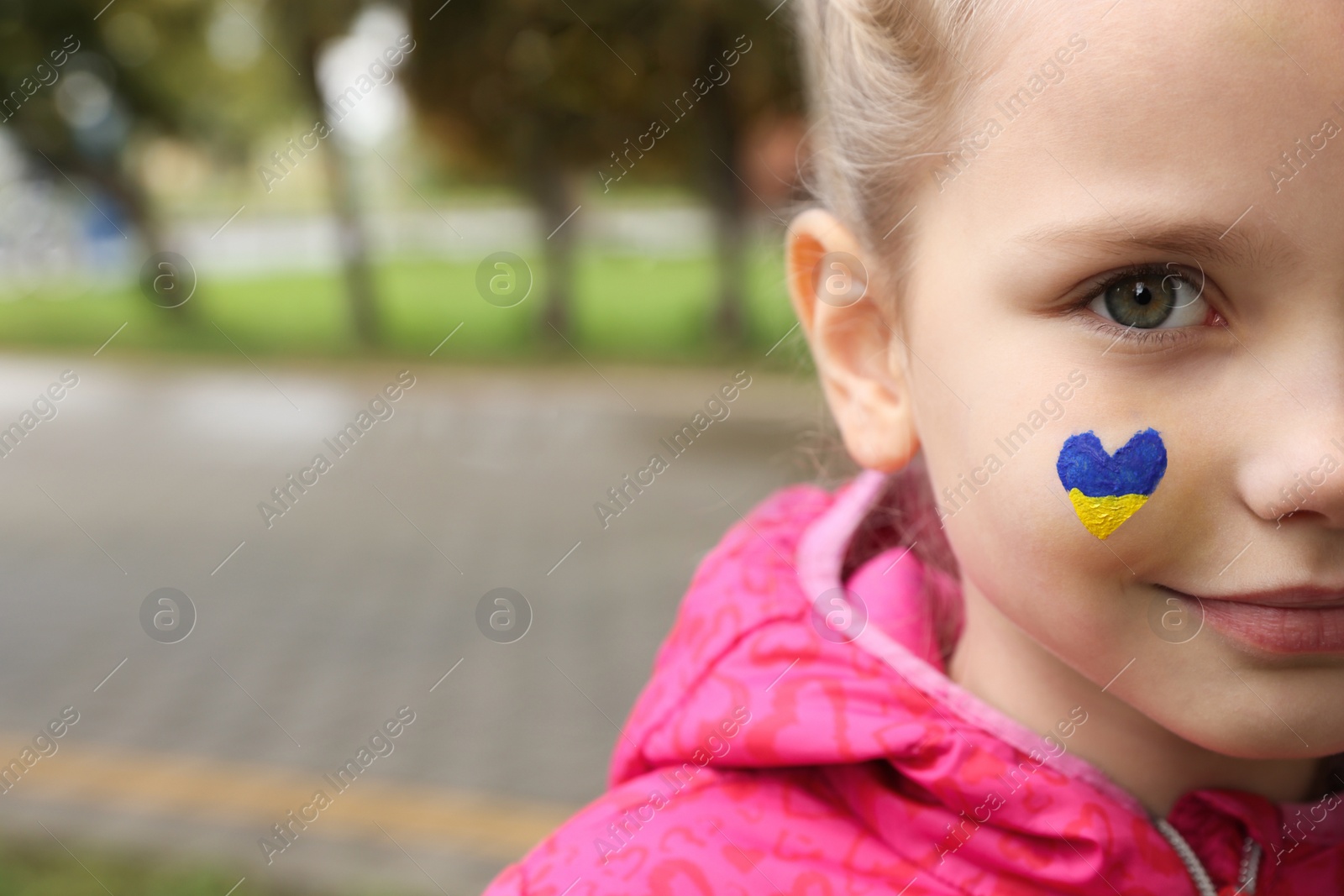 Photo of Little girl with drawing of Ukrainian flag on face in heart shape outdoors, closeup. Space for text