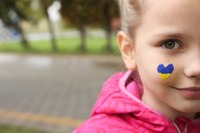 Photo of Little girl with drawing of Ukrainian flag on face in heart shape outdoors, closeup. Space for text