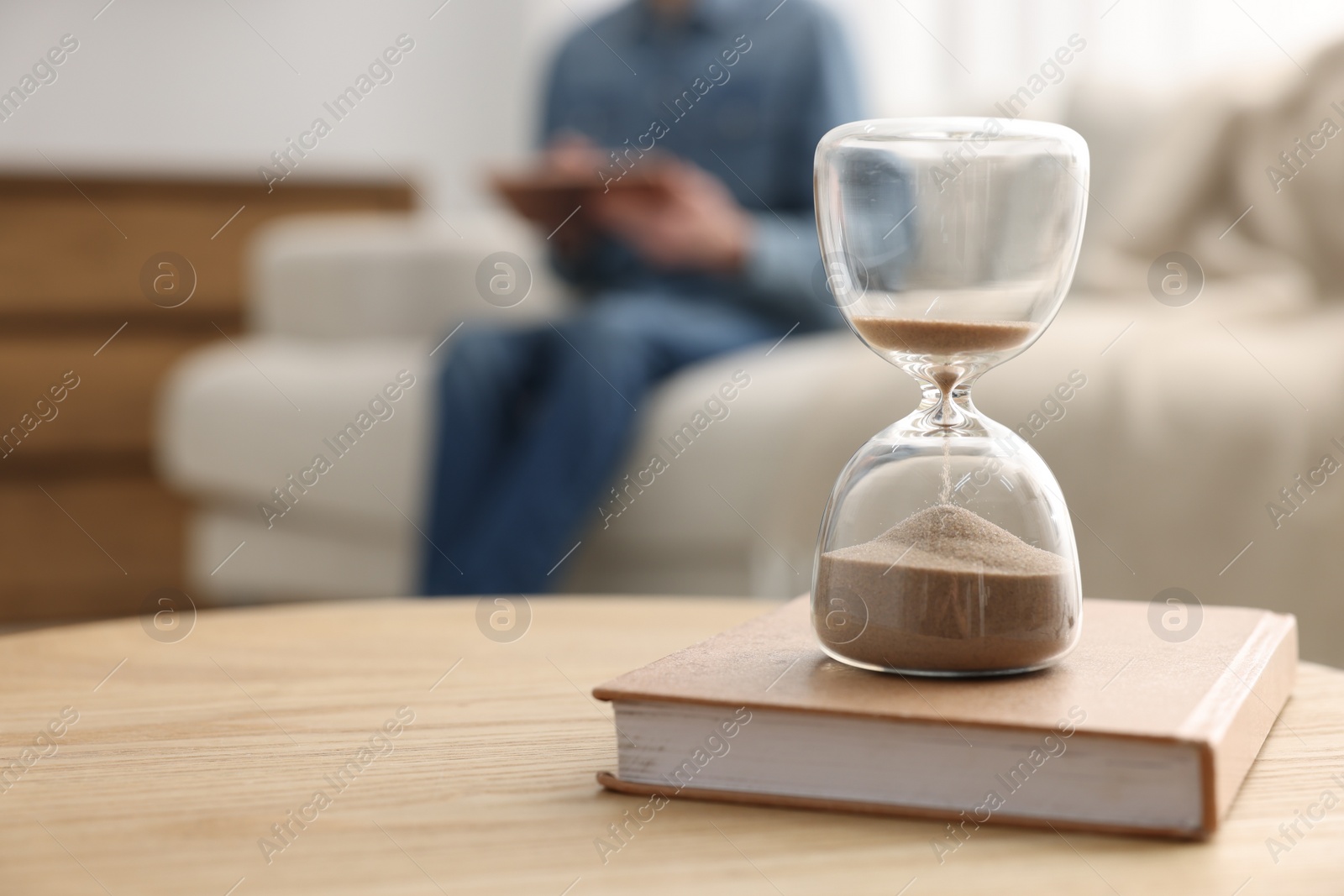 Photo of Hourglass with flowing sand on desk. Man reading book in room, selective focus