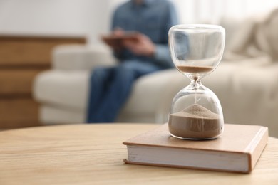 Photo of Hourglass with flowing sand on desk. Man reading book in room, selective focus