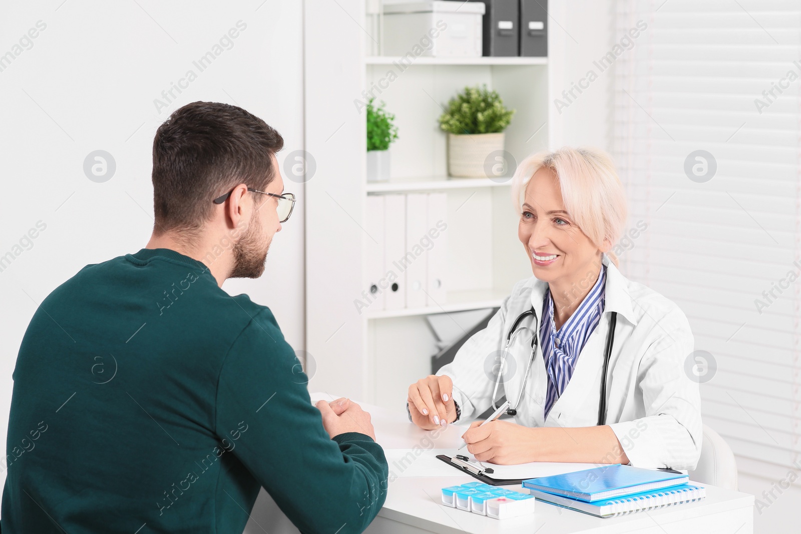 Photo of Doctor with pen and clipboard consulting patient at table in clinic