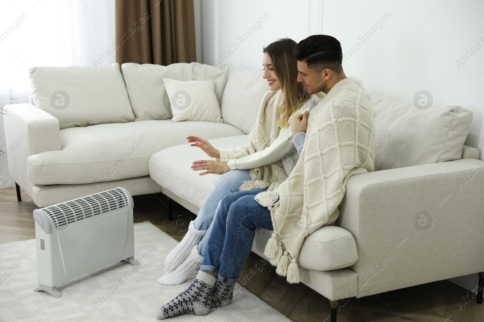 Photo of Happy couple sitting on sofa near electric heater at home