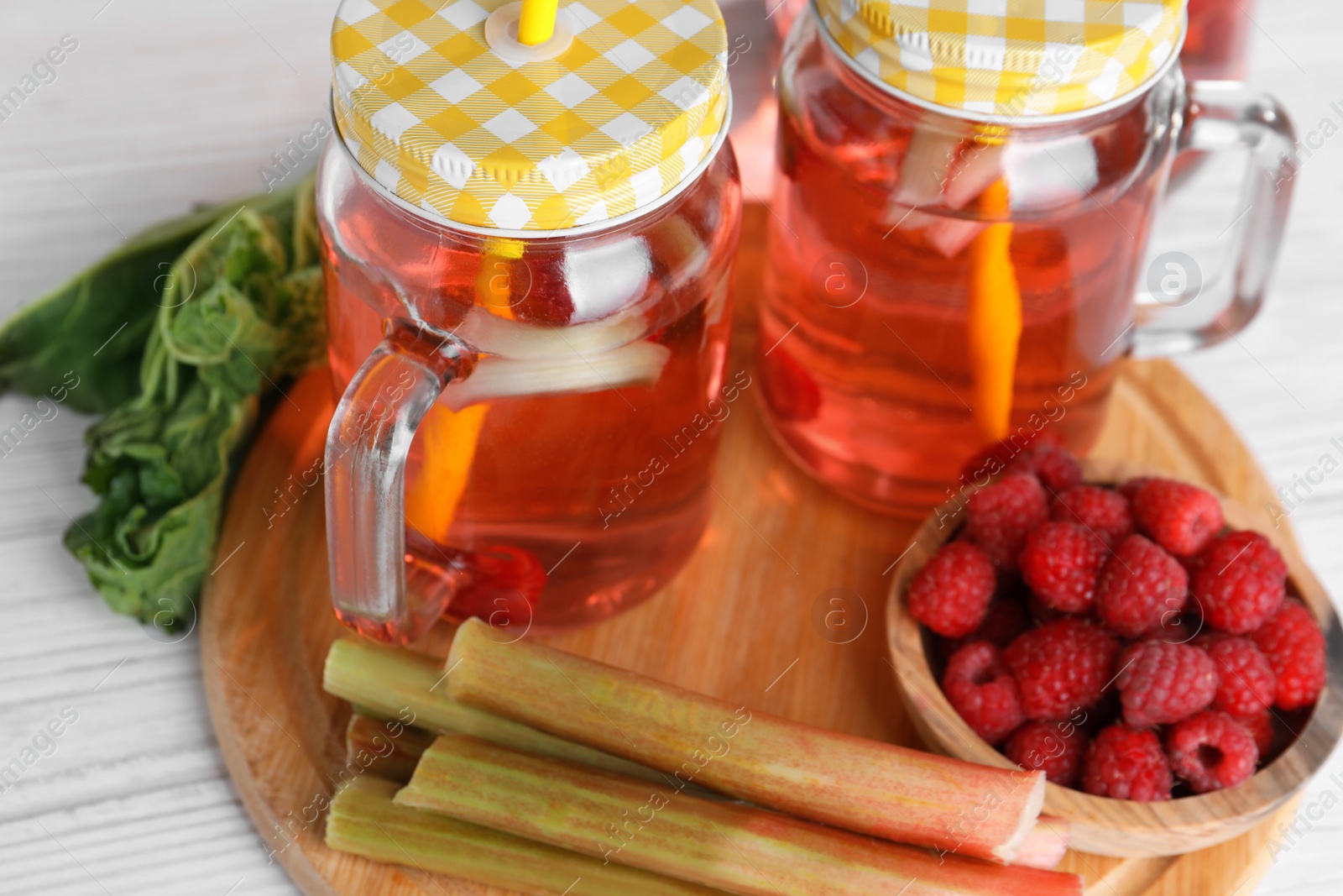 Photo of Mason jars of tasty rhubarb cocktail with raspberry and stalks on white wooden table