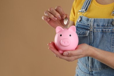 Photo of Woman putting coin into pink piggy bank on light brown background, closeup. Space for text