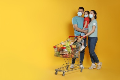 Photo of Family with protective masks and shopping cart full of groceries on yellow background. Space for text
